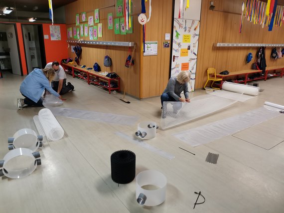 Drei Personen arbeiten an Kunststoffmaterialien auf dem Boden eines bunt dekorierten Raumes mit Holzverkleidung.