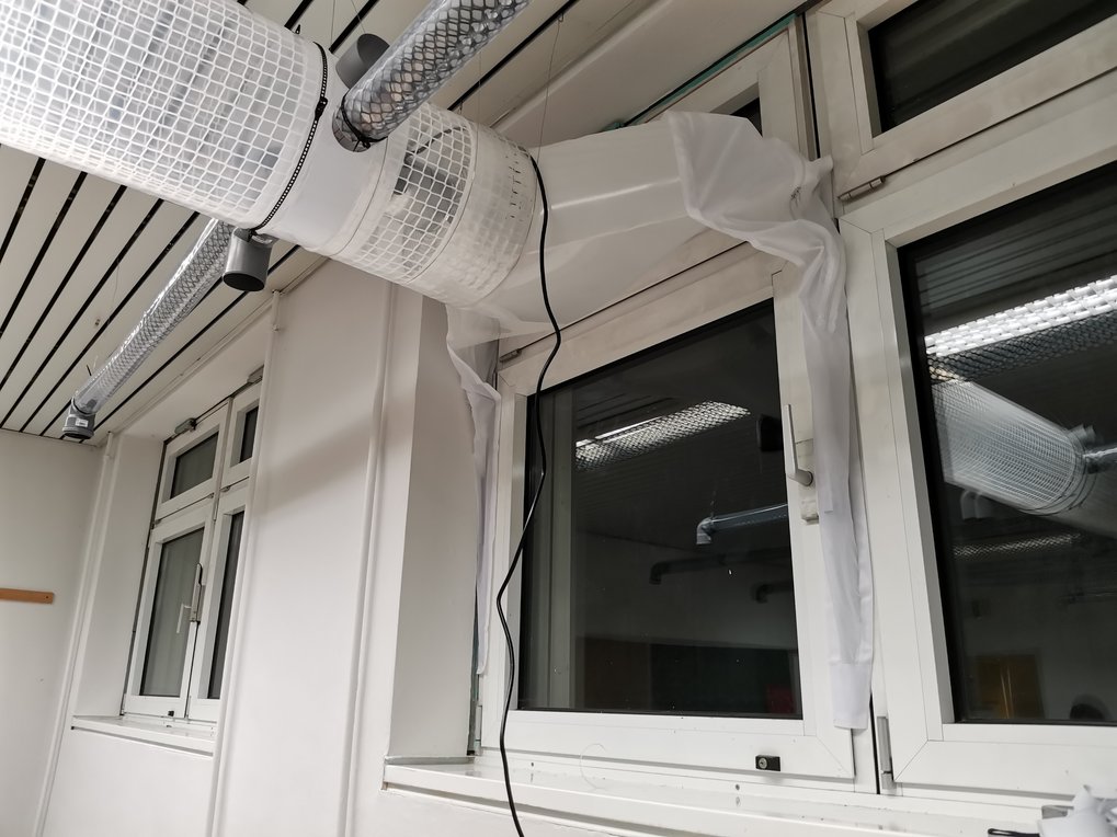 Raum mit weißem Lüftungsrohrsystem aus Kunststoff, über einem Fenster montiert. Ein Kabel hängt von der Installation.