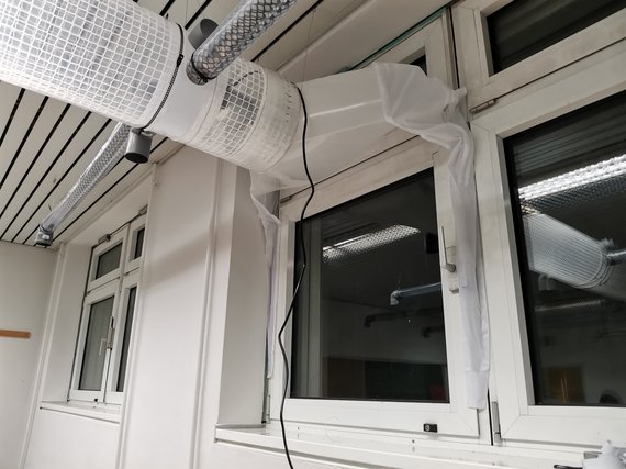 Raum mit weißem Lüftungsrohrsystem aus Kunststoff, über einem Fenster montiert. Ein Kabel hängt von der Installation.