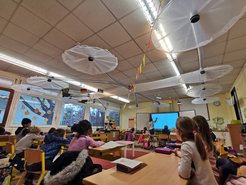 Im Klassenzimmer sitzen Schüler an Tischen, die Lehrkraft zeigt etwas auf der digitalen Tafel. Die Fenster sind mit Herbstmotiven dekoriert. Große Deckenventilatoren sind sichtbar.