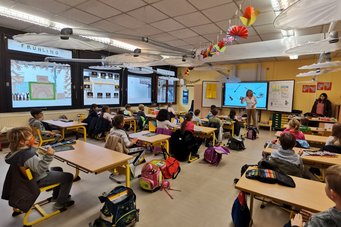 Die Brunnenschule in Mainz-Marienborn hat bereit elf Klassenzimmer mit dem Lüftungssystem ausgestattet. In einem gut beleuchteten Klassenzimmer sitzen Schüler an gelben Tischen, während zwei Lehrer eine Unterrichtsstunde halten. Dekorationen hängen von der Decke, und am Fenster sind Frühlingsmotive zu sehen. Schultaschen sind neben den Tischen platziert.