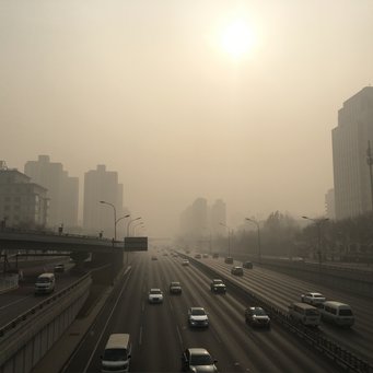 Smog über Peking. Dichte Smogschicht über einer Großstadtlandschaft (Peking, China), bei der Hochhäuser kaum sichtbar sind. Eine Autobahn mit geringem Verkehrsaufkommen erstreckt sich in die Ferne, während die Sonne schwach durch den Dunst scheint.