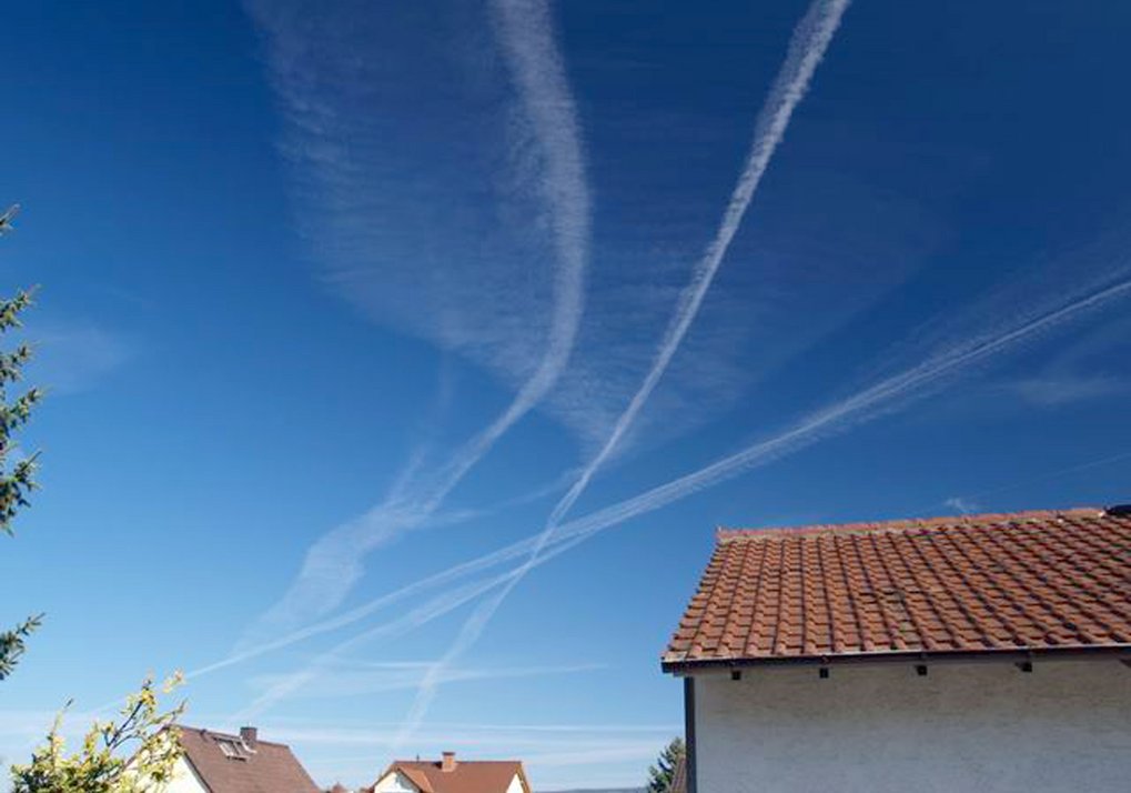 Die Eiskristalle der Kondensstreifen können bei feucht-kalten Bedingungen in Höhen von etwa 8 bis 12 Kilometern mehrere Stunden bestehen und hohe Wolken, sogenannte Kondensstreifen-Zirren bilden. Diese Wolken können je nach Sonnenstand und Untergrund lokal eine wärmende oder kühlende Wirkung entfalten. Blauer Himmel, Kondensstreifen, Häuserdächer