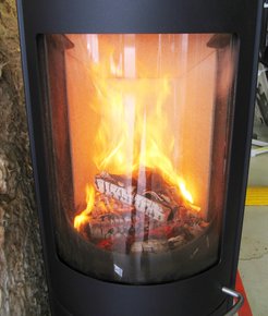 Field studies show that heating with private wood-burning stoves increases the concentration of fine dust in the immediate vicinity. Illustration of a private wood-burning stove, glass pane, stacked wood, flames (orange).