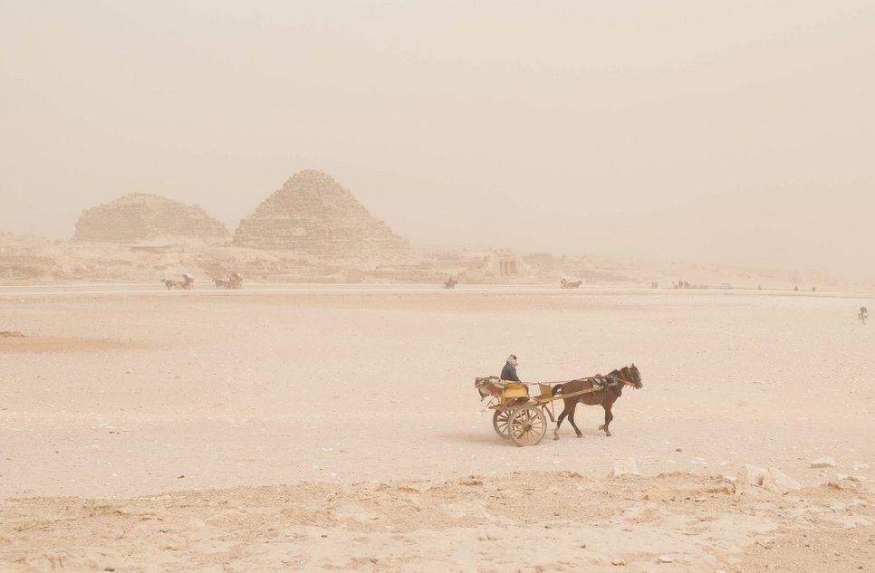 A horse-drawn cart traverses a sandy area with distant pyramids, under a hazy sky.
