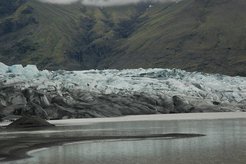 Eisiger Gletscher mit grauem Geröll erstreckt sich vor bewaldetem Berghang und ruhigem Wasser.