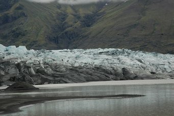 A new study suggests that a million years ago, glaciers began sticking more persistently to their beds, triggering cycles of longer ice ages. Glacier with ice and debris, surrounded by water, against a backdrop of green mountains.