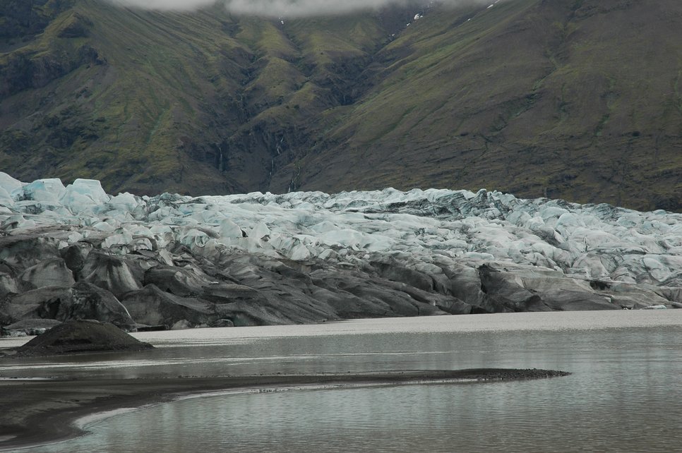 Glacier with ice and debris, surrounded by water, against a backdrop of green mountains.