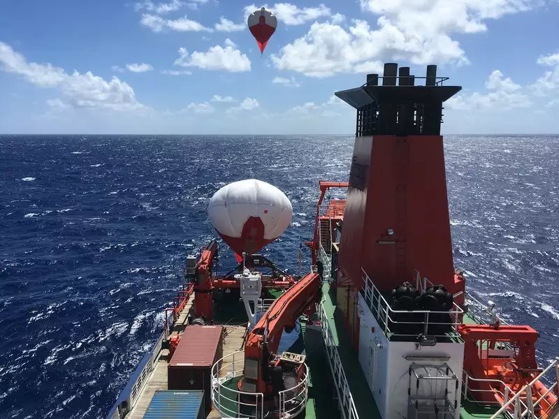 Ein rotes Forschungsschiff auf dem Ozean, im Hintergrund schwebt ein Ballon.