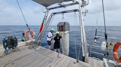 Two researchers on a boat deck are handling oceanographic sampling equipment, including a large net suspended above the sea, under a cloudy sky.