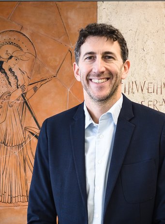 Eric Kort Mann in dunklem Sakko vor Wand mit antiker griechischer Zeichnung und Text.