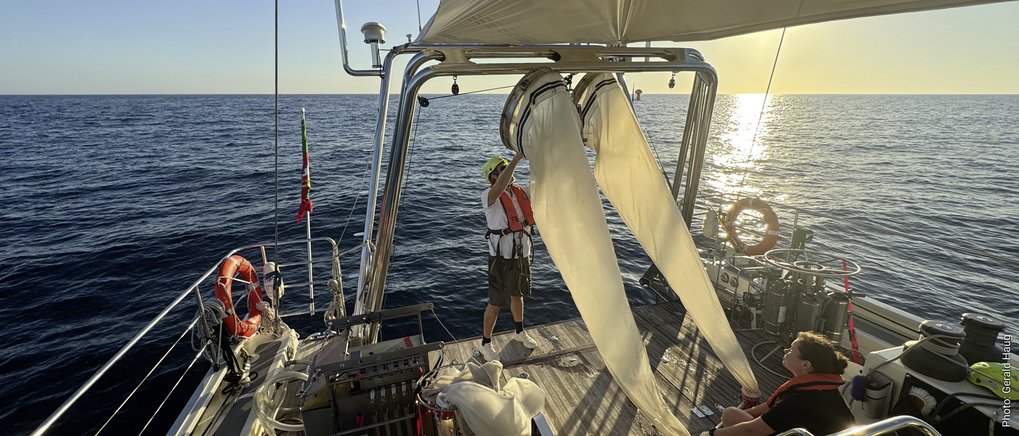Zwei Personen auf dem Deck eines Segelbootes bereiten Segel bei Sonnenuntergang auf ruhiger See vor.