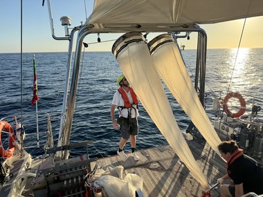 Auf einem Forschungsschiff im Meer sind zwei große Probenbehälter befestigt, daneben stehen zwei Personen mit Sicherheitsausrüstung.