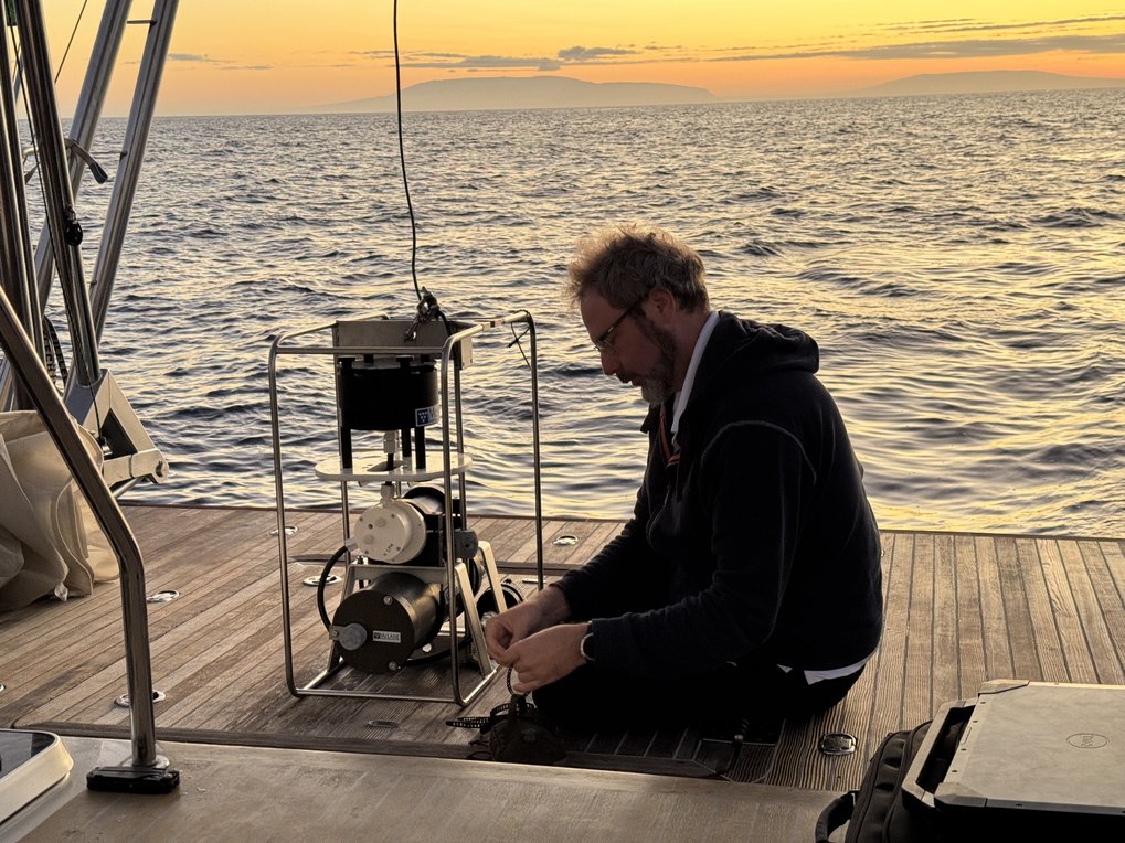 Forscher arbeitet auf einem Bootdeck mit einem technischen Messgerät, Meer und Sonnenuntergang im Hintergrund.
