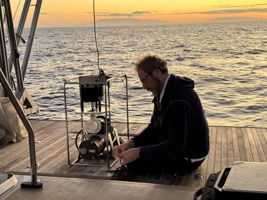 Forscher arbeitet auf einem Bootdeck mit einem technischen Messgerät, Meer und Sonnenuntergang im Hintergrund.