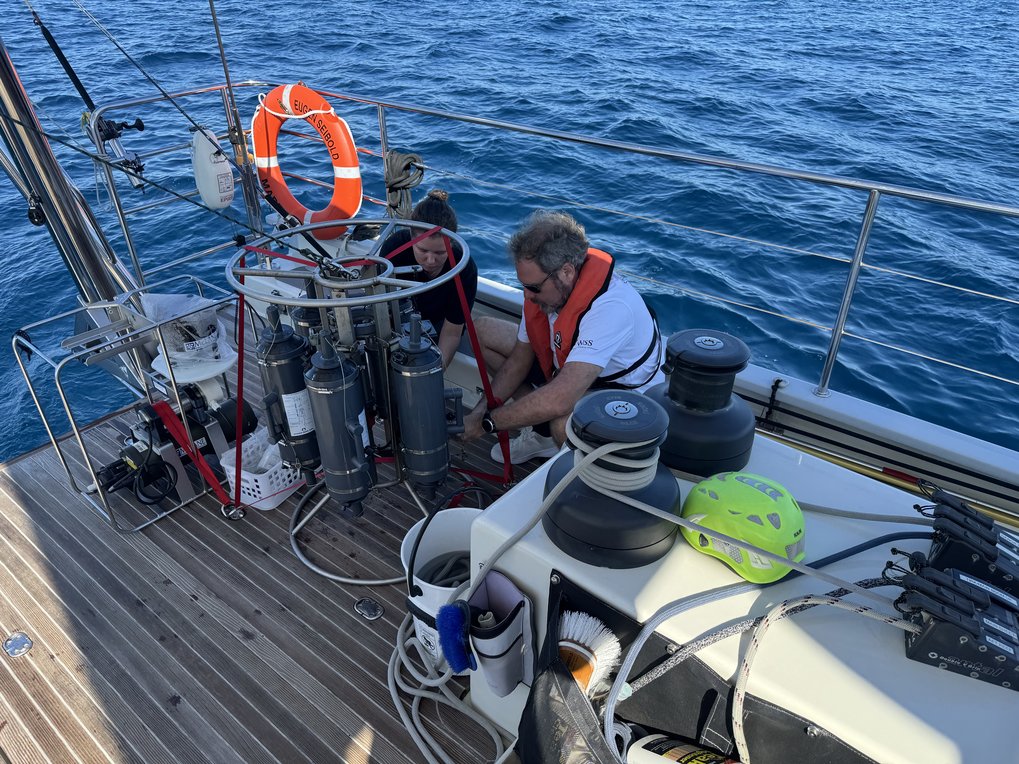 Zwei Personen mit Schwimmwesten arbeiten an einem wissenschaftlichen Gerät auf einem Segelboot.