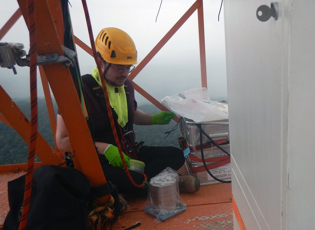 Person in Schutzkleidung kontrolliert Ausrüstung auf einem orangefarbenen Metallgerüst in großer Höhe.